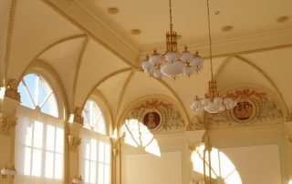 Eleganter Festsaal mit hohen Rundbogenfenstern, verzierten Wandmalereien und dekorativen Kronleuchtern an einer cremefarbenen Decke.