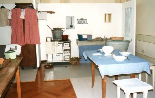 Historische Kücheneinrichtung mit Herd, Waschbecken, Tisch mit blau-karierter Tischdecke und Hockern sowie hängender Kleidung in einem Ausstellungsraum.