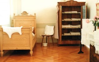 Schlafzimmer mit einem hölzernen Bett, einem offenen Schrank mit gefalteter Bettwäsche und einem Stuhl.