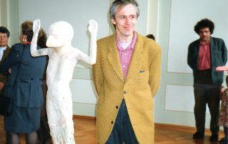 Person in gelbem Sakko neben einer abstrakten Skulptur, im Hintergrund weitere Personen in einem Ausstellungsraum.