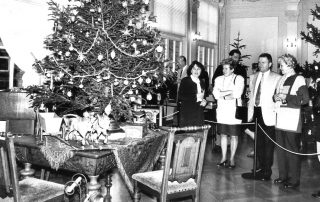Historisches Schwarz-Weiß-Foto einer Weihnachtsausstellung mit geschmücktem Weihnachtsbaum, antikem Tisch, Spielzeugpferden und einer Gruppe von Personen, die die Ausstellung betrachten.