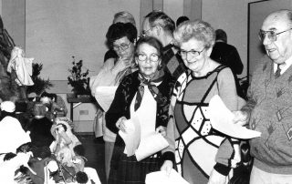 Schwarz-Weiß-Foto einer Gruppe älterer Personen, die eine Ausstellung mit handgefertigten Figuren und Dekorationen betrachten, einige halten Papiere in den Händen.