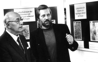 Zwei Männern vor Ausstellungstafeln mit Luftaufnahmen und Beschreibungen zum Zweiten Weltkrieg.