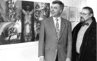 Zwei Männern vor einer Ausstellung mit expressiven Gemälden, darunter eine Darstellung von Jesus am Kreuz.