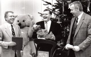 Schwarz-Weiß-Foto von drei Männern in Anzügen, lachend vor einem geschmückten Weihnachtsbaum, einer hält einen Teddybären in den Händen.