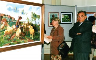 Besucher einer Kunstausstellung betrachten ein Gemälde eines Schäfers mit Schafen in einer Berglandschaft.