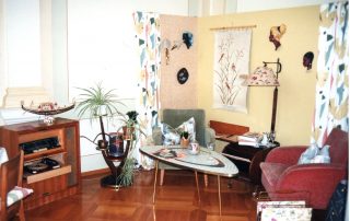 Historisches Wohnzimmer mit Retro-Möbeln, darunter ein Radiogramm, ein Couchtisch mit Dekorationen, zwei Sesseln, Zimmerpflanzen und Wanddekorationen.